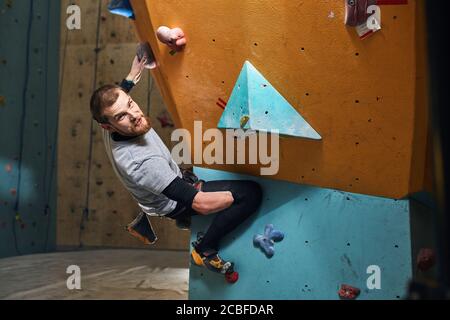Junger gutaussehender, körperlich behinderter Boulderer mit Bart, der an einer farbenfrohen Kletterwand hing, hat gerade sein aktives Training beendet und dabei den Witz der Kamera betrachtet Stockfoto
