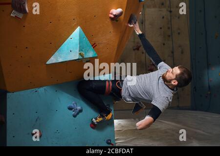 Seitenansicht des motivierten starken Kletterers mit körperlicher Behinderung, Training an bunten künstlichen Kletterwänden im Bouldersportzentrum, overh Stockfoto