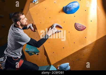 Junge ehrgeizige körperlich behinderte Mann Training in Boulderhalle, Klettern auf künstlichen bunten Felswand, tragen Geschirr und Kreidebeutel, hat eine Stockfoto