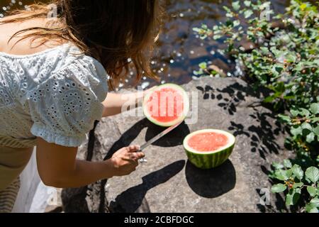 Von hinten von einer Frau in weiß gekleidet, die saftige, reife Wassermelone in zwei auf dem Felsen beim Bett des Baches oder des Flusses aufschneiden. Kanadischer Sommer. Stockfoto