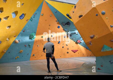 Junger männlicher Boulderer mit körperlicher Behinderung, der nachdenklich in der Kletterhalle steht, bunte künstliche Felswände anschaut und wählt, wo er steht Stockfoto