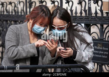 Zwei asiatische Frauen, die Gesichtsmasken tragen, schauen sich ihre Handys außerhalb des Hofes an. London, England Stockfoto