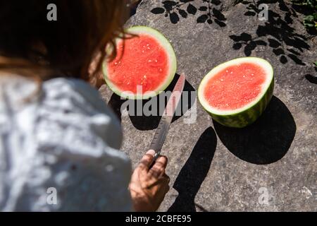 Nahaufnahme von hinten von einer Frau in weiß gekleidet, Schneiden eine saftige, reife Wassermelone in zwei auf einem Felsen am Bett eines Baches oder Flusses. Stockfoto