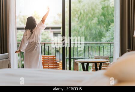 Glückliche Frau streckt und öffnet die Vorhänge am Fenster am Morgen. Süße Träume, neuer Tag, Wochenende, Urlaub Konzept Stockfoto