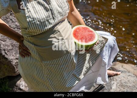 Nahaufnahme einer schwangeren Frau, die eine halbe, saftige und rote Wassermelone auf ihrem Schoß neben ihrem großen Bauch hält. Bach oder Fluss im Hintergrund. Stockfoto