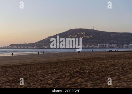 Agadir, Marokko Strand mit marokkanischem Motto auf dem Berg. Schreiben auf dem Hügel Bedeutung, Gott, Land, König. Abenduntergang mit Leuten, die weiter gehen Stockfoto