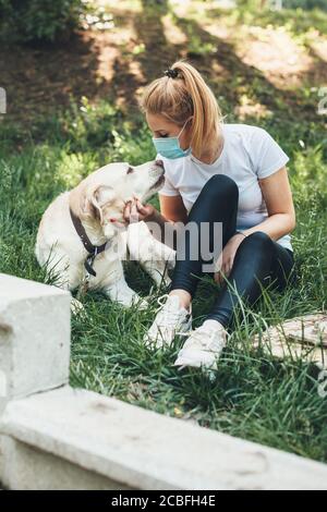 Vorderansicht Foto von einer blonden Frau trägt einen Anti Virusmaske, die mit ihrem labrador auf dem Gras liegt Stockfoto