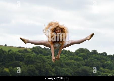 Eine junge Ballerina, die ein weißes Tutu trägt, macht einen Spaltsprung in der Luft, ihre langen blonden Haare fliegen nach oben. Stockfoto