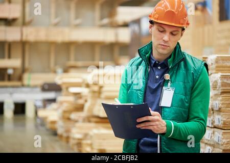 Junger Mann in Uniform und Helm, Lagerarbeiter vergleicht Produktdaten, liest ein Dokument und nimmt Notizen auf dem Markt Stockfoto