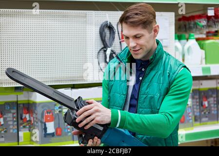 Junge kaukasische Verkäufer in grünen Uniform halten Ausrüstung in den Händen, elektrische Säge zu einem attraktiven Preis auf dem Markt, Mann prüft es Stockfoto