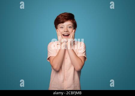 Überrascht kaukasischen Jungen mit roten Haaren bedeckt Wangen mit Handflächen Lächelt auf einer blauen Studiowand Stockfoto