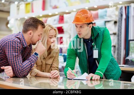 Selbstbewusster junger Arbeiter des Vertriebszentrums erklären den Kunden, Lagerarbeiter im Helm zeigen auf Dokument und erklären die Anweisung, schöne Co Stockfoto