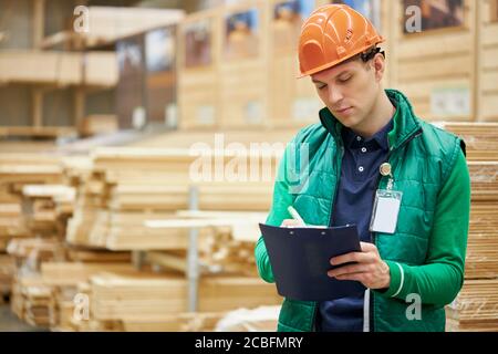 Konzentrierte selbstbewusste Lagerarbeiter schreiben die Anzahl der Bretter in einem Dokument, hart arbeitende männliche Arbeit in großen Verteilzentrum. Viel Holz Stockfoto