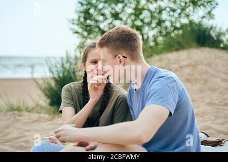 Junges Paar sitzt am Strand und füttert sich gegenseitig Stockfoto