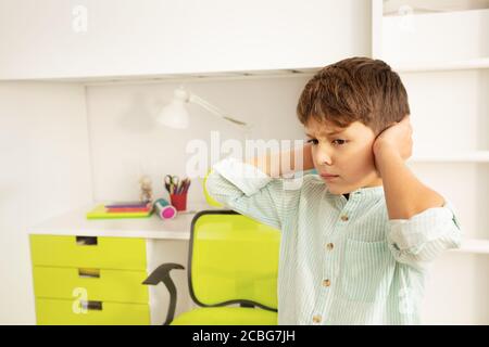 Kleiner autistischer Junge mit trauriger negativer Gesichtsausdruck, der die Ohren schließt Und grinst die Zähne Stockfoto