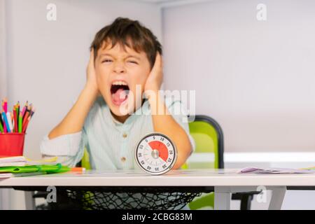 Trauriger schreiender autistischer Junge Portrait nahe Ohren mit Händen während der Übung aba Therapiekurs mit Stundenzähler, Hörempfindlichkeitsproblem Stockfoto