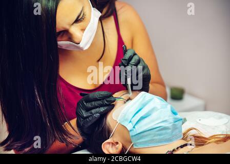 Frau setzen falsche Wimpern, um Dame im Schönheitssalon mit Masken, neue normale Stockfoto