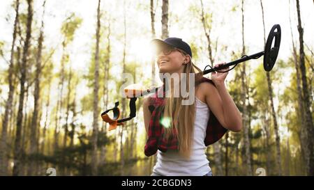 Erfolgreiche Suche. Junge Frau hält Metalldetektor im Wald. Direkte Sonneneinstrahlung. Hochwertige Fotos Stockfoto