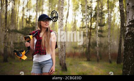 Eine junge Frau mit einem Lächeln hält einen Metalldetektor über ihre Schulter im Wald. Hochwertige Fotos Stockfoto