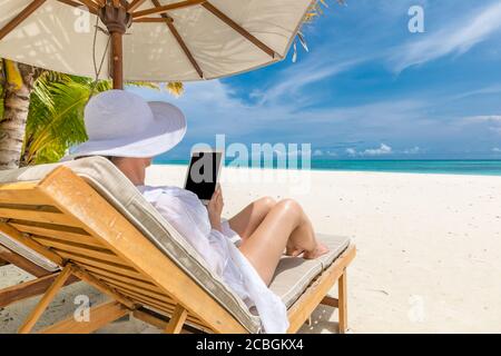 Frau mit Tablet-Computer am Strand sitzt in der Lounge Liegestuhl. Junge Frau Hut mit Tablet-pc am Strand, Luxus Sommerurlaub, Freizeit Stockfoto