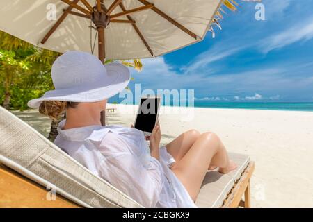 Frau mit Tablet-Computer am Strand sitzt in der Lounge Liegestuhl. Junge Frau Hut mit Tablet-pc am Strand, Luxus Sommerurlaub, Freizeit Stockfoto