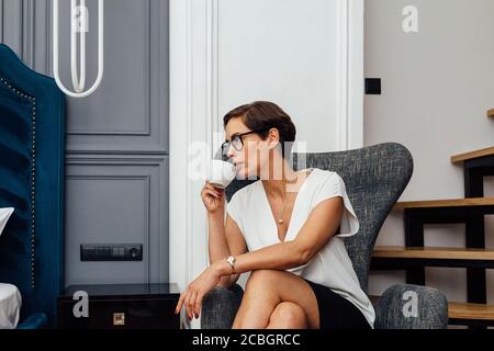 Seitenansicht einer Frau mittleren Erwachsenen, die Kaffee trinkt in einem modernen Apartment. Geschäftsfrau in einem Hotelzimmer, die in einem Sessel sitzt und wegschaut. Stockfoto