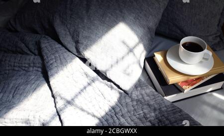 Morgenlicht auf grauem Bett mit einem Stapel Bücher Und eine Tasse frischen Kaffee Stockfoto