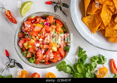 Tex Mex Pico de Gallo Tomatensalsa mit Nacho-Tortilla-Chips. Die Schüssel ist von oben gesehen, flach gelegt, und es gibt Zutaten verstreut auf dem Tisch: Stockfoto