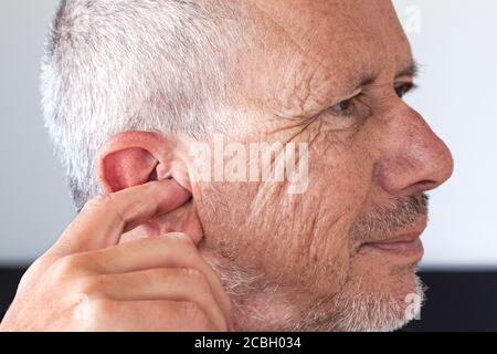 Älterer Mann mit unrasiertem Bartstoppel, der sein Ohr mit dem Zeigefinger pflückt. Nahaufnahme des Bildes. Stockfoto