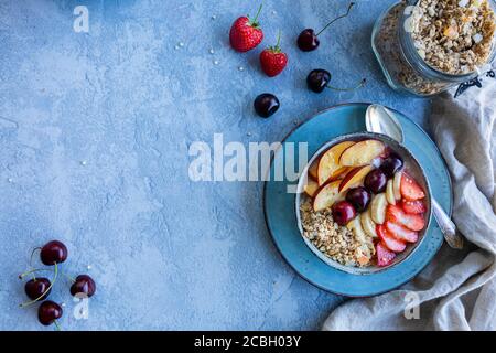 Eine Schüssel mit frischem Obst, Müsli und Joghurt. Gesundes Frühstückskonzept. Die blaugrauen Schalen sind mit Erdbeeren, Pfirsichen, Banane und cherri gefüllt Stockfoto