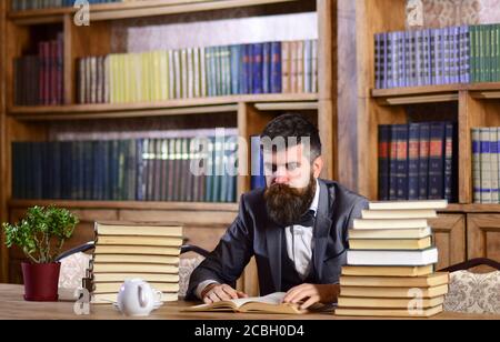 Wissenschaft und Studium Konzept. Mann im klassischen Anzug oder Professor mit konzentriertem Gesicht sitzt in der Bibliothek neben Haufen von Büchern, Bücherregale auf dem Hintergrund, entkokelt. Forscher oder Lehrer Buch lesen. Stockfoto