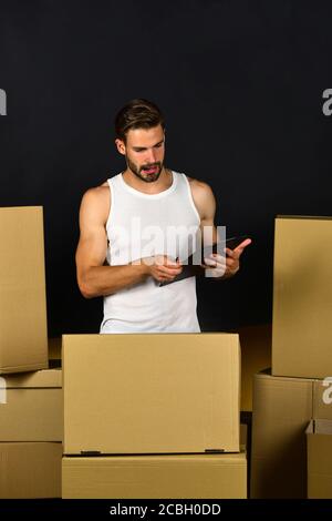 Macho mit Bart und konzentriertem Gesicht. Kerl mit Clip-Board und Stift isoliert auf schwarzem Hintergrund. Lieferung und Umzug im Konzept. Mann mit starken Armen, der zwischen Kartons steht. Stockfoto