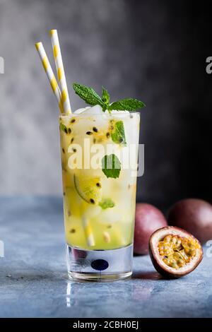 Passionsfrucht Mojito Cocktail und Mocktail in einem hohen Glas, mit zerstoßenem Eis, Limette und Minzblättern. Passionsfrüchte auf dem Tisch. Grauer Hintergrund mit Stockfoto