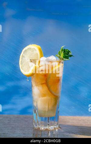 Ein Glas Eistee mit Zitrone und Minze, am Pool mit blauem Wasser im Hintergrund. Vertikales Bild. Am Rand des Glases A befindet sich eine in Scheiben geschnittene Zitrone Stockfoto