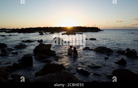 Ein Paar sitzt im Meer und genießt den Sonnenuntergang an der North Shore, Hawaii. Stockfoto