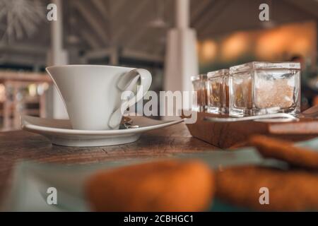 Niedriger Blick auf Holztisch mit weißem Kaffeebecher, braunem Zucker und Keksen. Entspannender Coffee Shop in der Nähe, Morgenlicht, positive Vibes Stockfoto