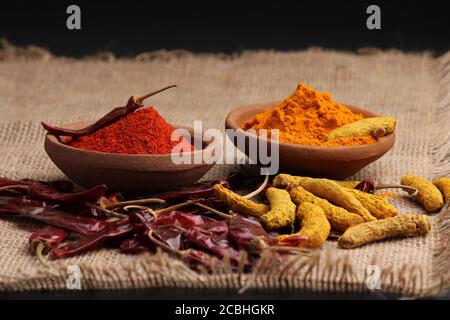 Indische Gewürze Chili und Kurkuma Pulver in einem Tontopf mit roten Chilis und Kurkuma Wurzel auf Jute Hintergrund. Stockfoto