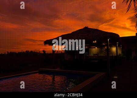 Schöner orange feuriger Sonnenuntergang in den Tropen. Straßendach von Heu, neben dem Pool und mit Blick auf das Reisfeld Stockfoto