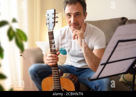 Lächelnder Mann mit seiner Akustikgitarre in der Hand und geradlinig Vorne auf dem Sofa sitzend mit Musikständer mit Liedern Zu Hause Stockfoto