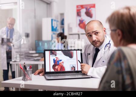 Kardiologin im Gespräch mit einer älteren Patientin im Krankenhausbüro über ihre Herzdiagnostik und Genesungsbehandlung mit weißem Mantel und Stethoskop. Ältere Arzt schriftlich auf Zwischenablage im Klinikkorridor. Stockfoto