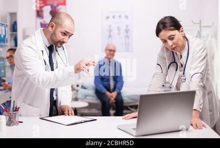 Ärzteteam überprüft die Diagnose des Patienten auf dem Laptop, während er besorgt auf dem Krankenhausbett sitzt. Älterer Mann während der Konsultation. Sanitäter mit Stethoskop. Stockfoto