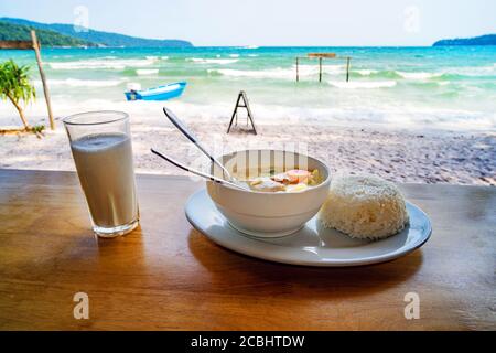 Thailändische Küche. Ein Teller Suppe mit Kokosmilch und gekochtem Reis auf dem Tisch vor der wunderschönen Meereslandschaft. Frühstück im Strandrestaurant Stockfoto