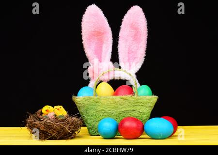 Vogelnest mit kleinen Spielzeugküken in der Nähe von Korb mit bunten handgefertigten Ostereiern und mehreren roten und blauen Eiern außerhalb davon auf gelben Tisch gelegt und isoliert auf schwarzem Hintergrund. Osterdekorationen Stockfoto