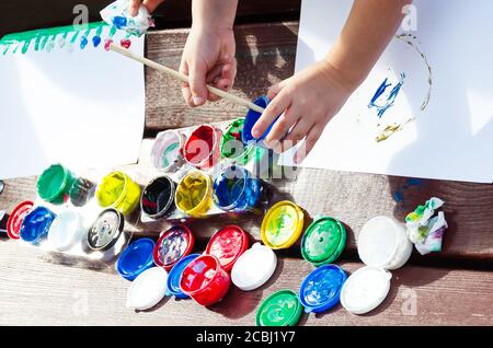 Kinderhände zeichnen Gouache Farben auf weißem Hintergrund. Offene Gläser mit mehrfarbiger Farbe. Pinsel von Hand in Farbe tauchen. Im Freien Stockfoto
