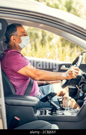 Profilansicht eines glücklichen älteren Mannes Mitte 60 mit einer medizinischen Maske während einer Epidemie. Taxifahrer in einer Maske, Schutz vor dem Virus. Alleinreisende älterer Mann am Steuer eines Autos. Stockfoto