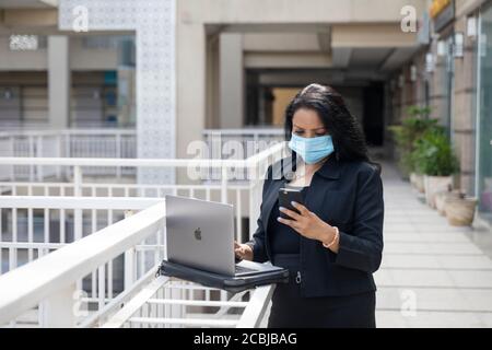 Eine indische Frau mit Covid-19 Schutzmaske arbeitet auf ihrem Laptop in einer städtischen Unternehmensumgebung, Stockfoto