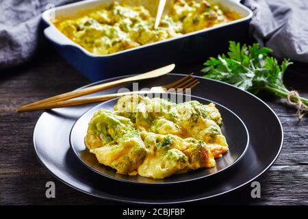 Eine Portion Cheesy Broccoli backen, Broccoli Auflauf auf einem schwarzen Teller auf einem Holztisch mit Backform im Hintergrund, horizontale Ansicht von abov Stockfoto