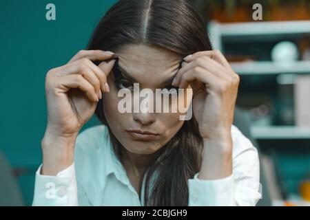 Müde schläfrige Geschäftsfrau versucht, nicht im Büro einschlafen, während klebt Klebeband auf ihre Augen und hält die Augen mit den Händen. Getöntes Bild Stockfoto