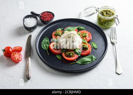 Burrata Büffelkäse serviert mit frischen Tomaten und Basilikumblättern Pesto-Sauce auf schwarzem Teller weißem Hintergrund Stockfoto
