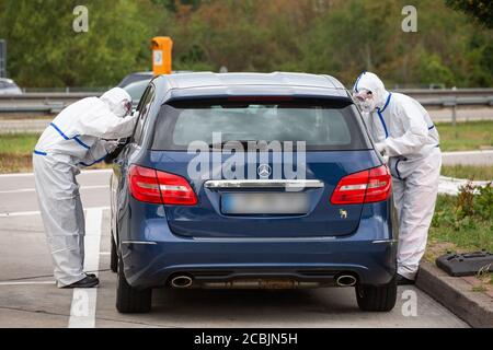 14. August 2020, Baden-Württemberg, Neuenburg am Rhein: Zwei Mitarbeiter des DRK stehen in einem Schutzanzug am offenen Fenster eines Autos, während sie den Abschmiertest für den Corona-Test auf einem Autobahnparkplatz durchführen. Nach der Rückkehr aus dem Urlaub können sich Reisende auf dem Parkplatz Neuchâtel-Ost auf das neuartige Corona-Virus testen lassen. Foto: Philipp von Ditfurth/dpa - ACHTUNG: Die Nummernschilder wurden aus persönlichen Gründen verpixelt. Stockfoto